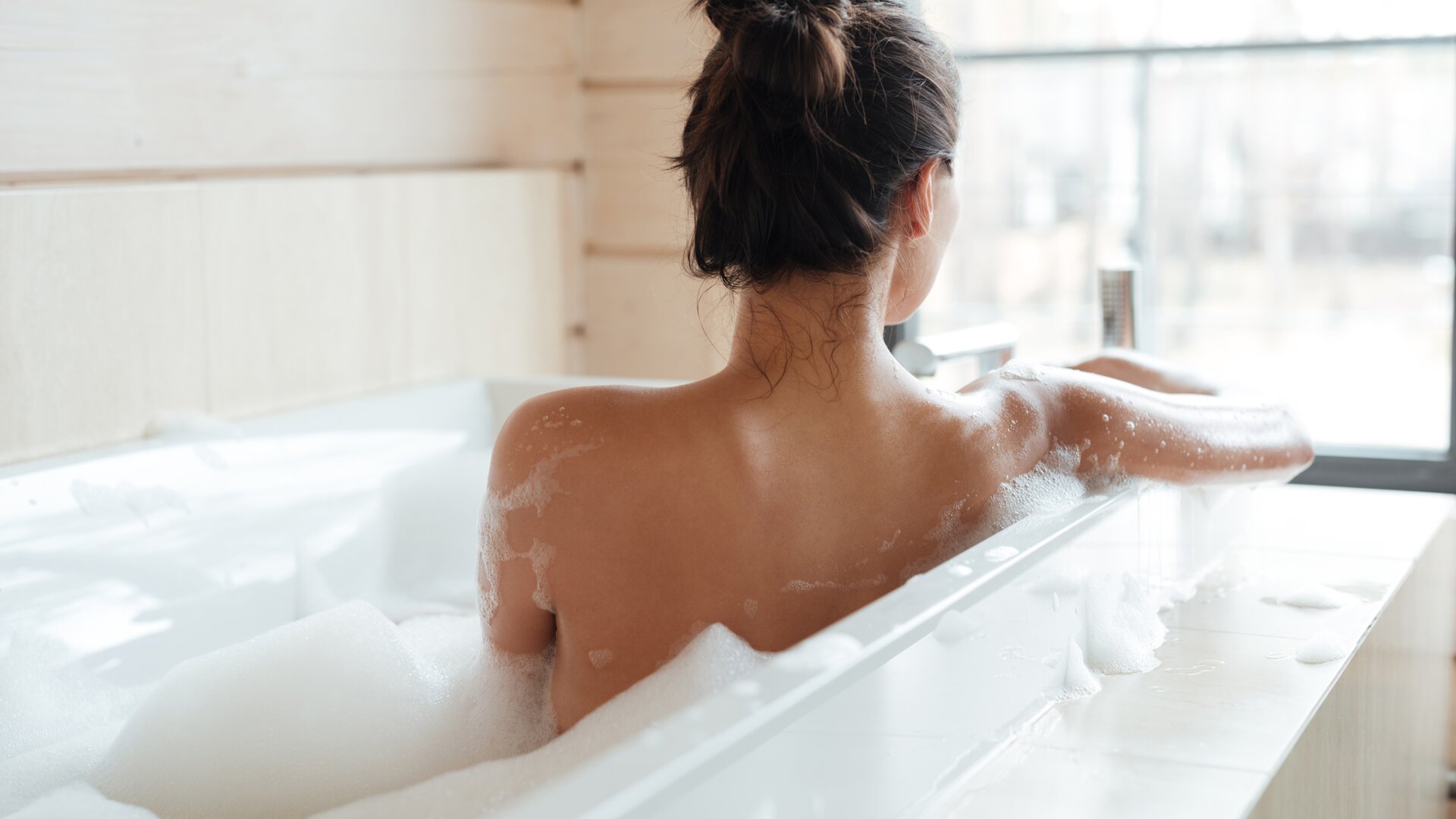 Schiena di una donna, seduta in una vasca da bagno bianca riempita di schiuma, che guarda la natura dalla finestra.